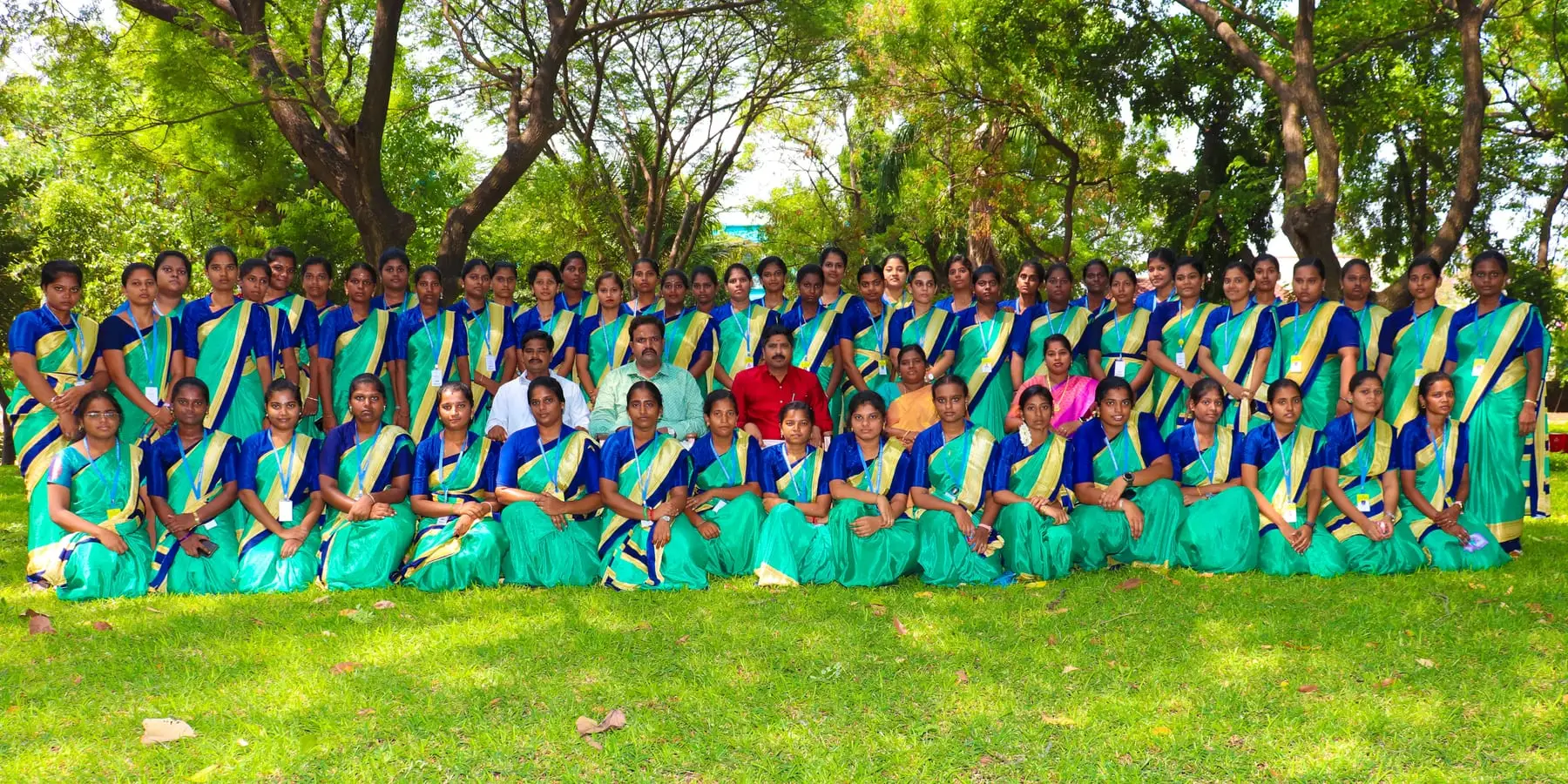 A batch of students in green and blue saris poses with faculty on a lawn.
