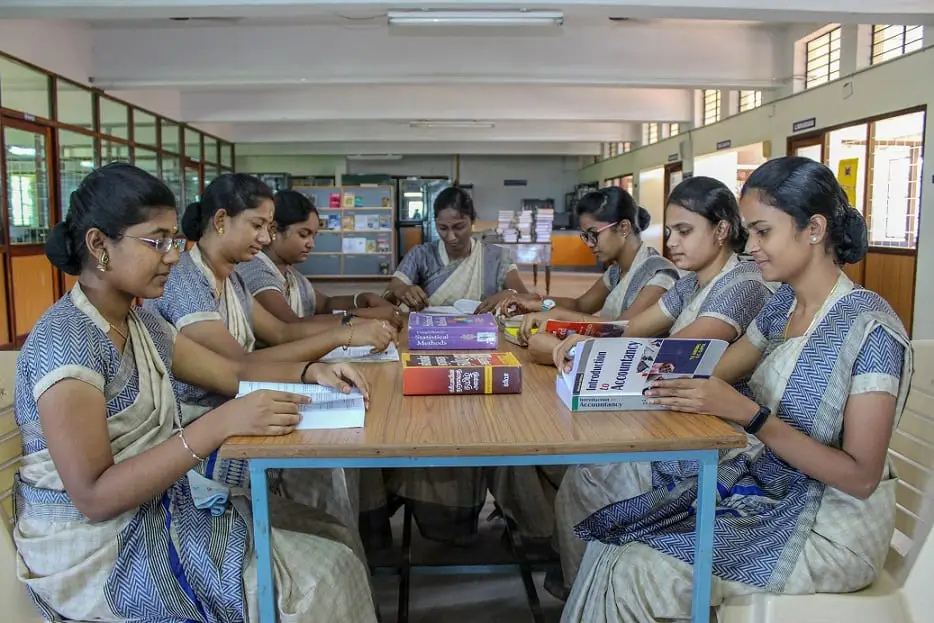 Student-teachers in uniform saris work in groups around tables in a bright classroom.