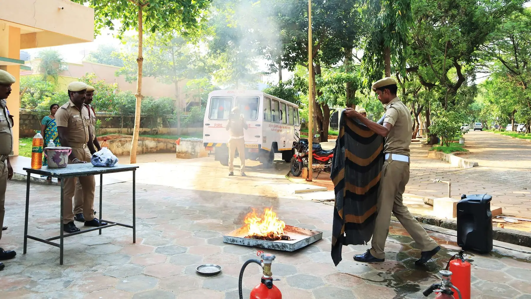 Fire service officers demonstrate fire fighting to students standing in a circle outdoors.