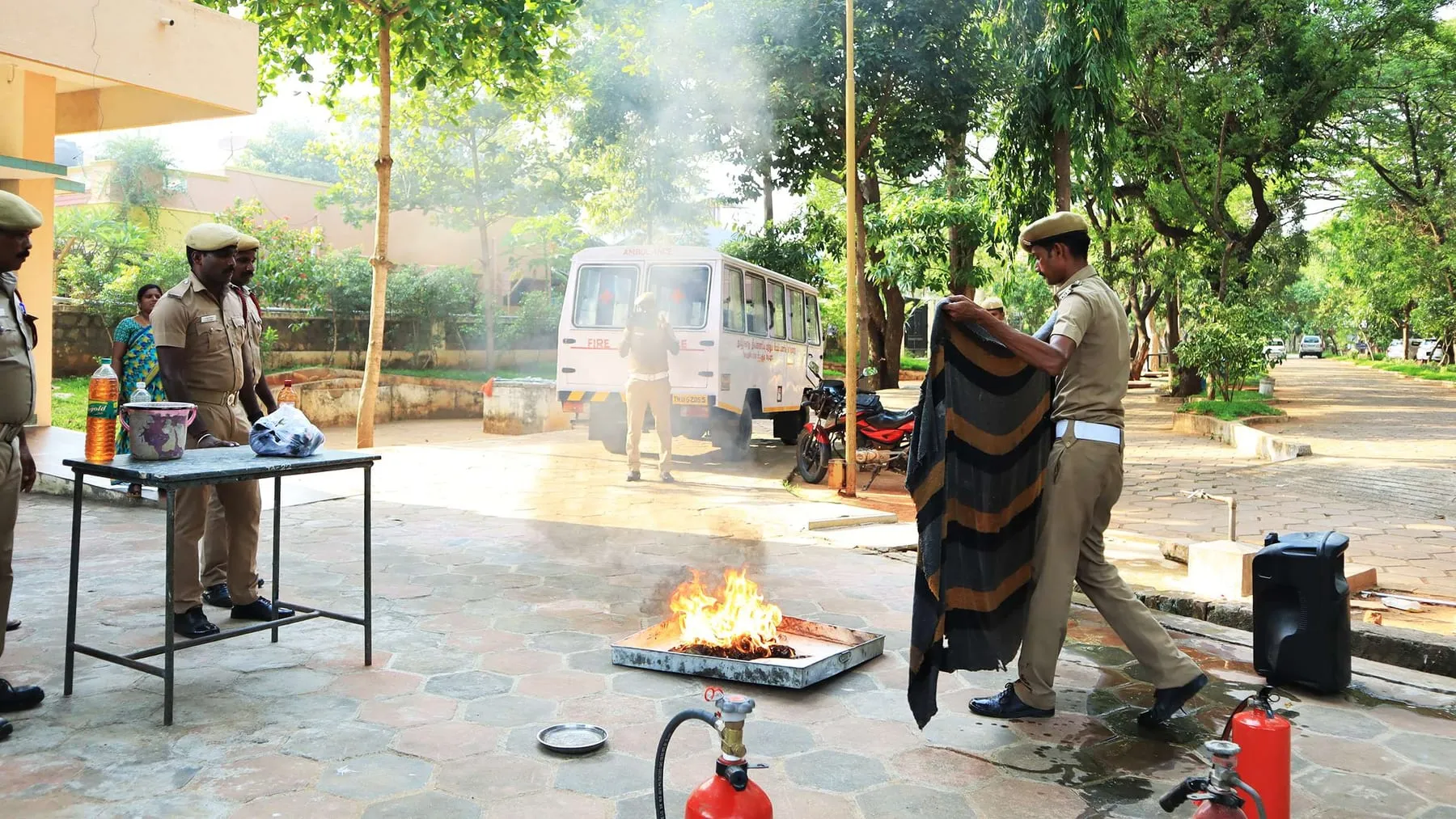 Fire service officers demonstrate fire fighting to students standing in a circle outdoors.