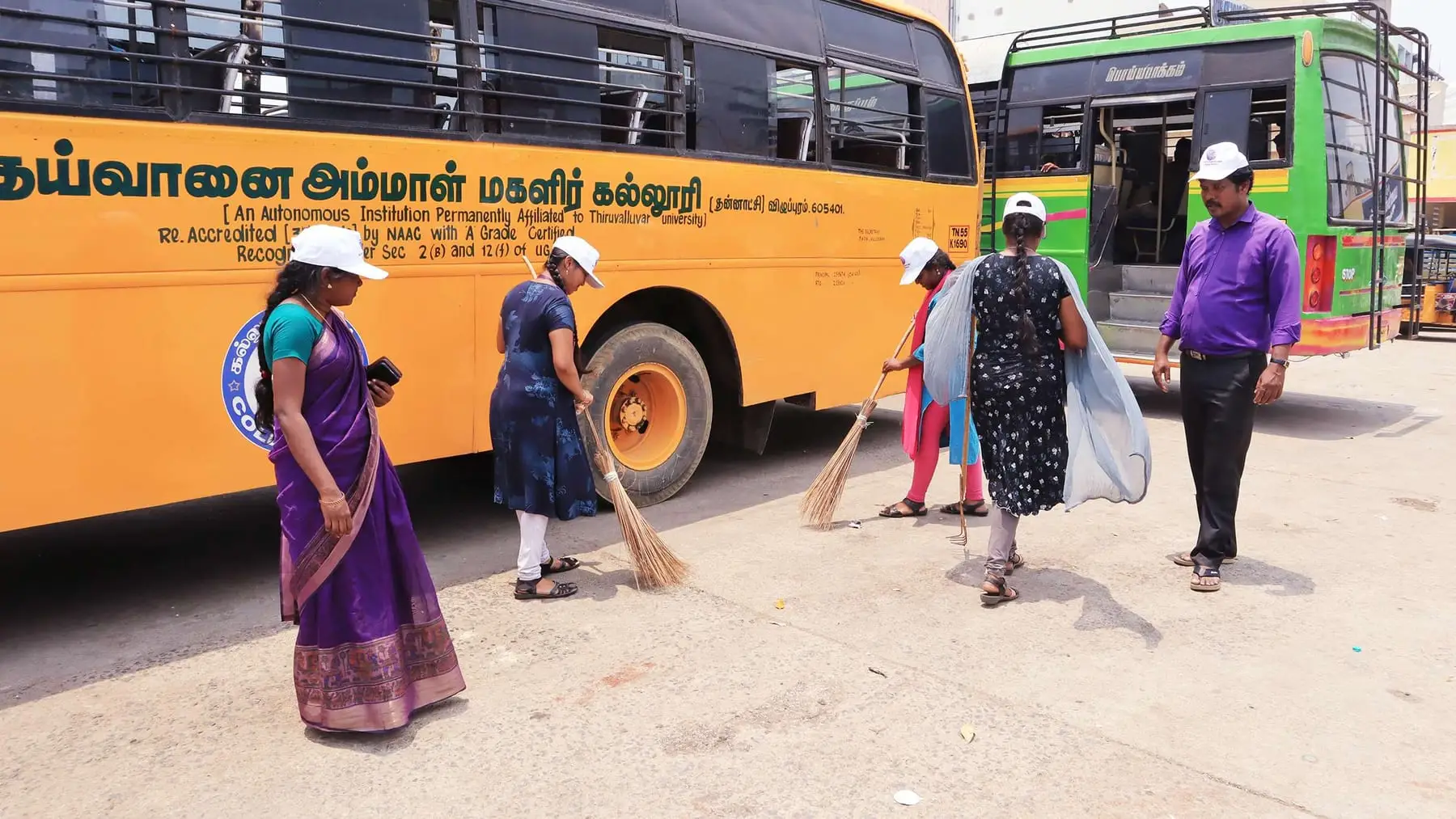 Students in white caps sweep the road next to parked buses.